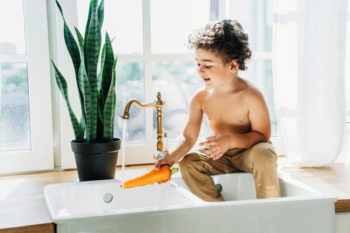 Happy caucasian curly boy sitting on sink at kitchen washing carrot under water. Handsome kid home